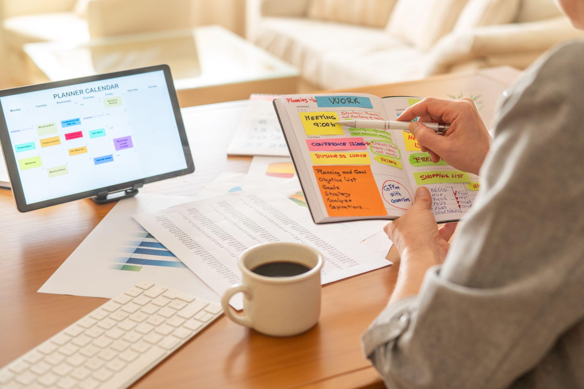 Person planning a content schedule in a notebook beside a tablet calendar and reports on a desk.