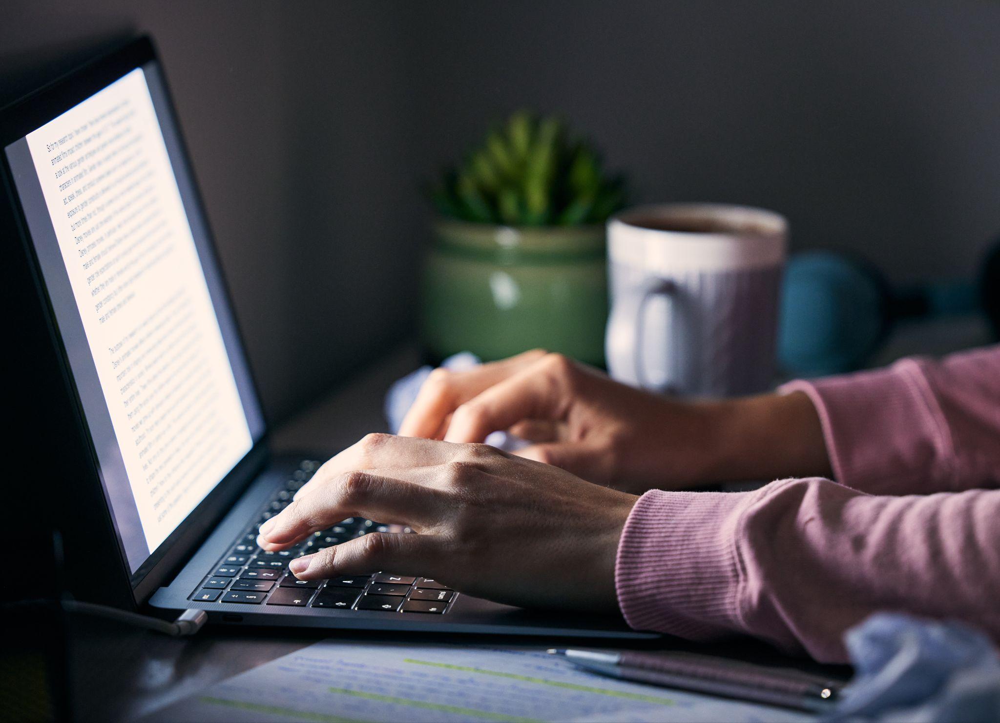 Person typing on a laptop at a desk at night with a coffee mug, plant, and notes nearby
