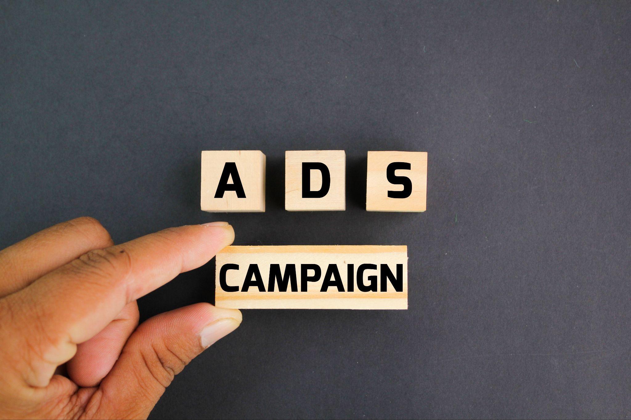 Hand arranging wooden blocks that spell Ads Campaign on a dark gray background